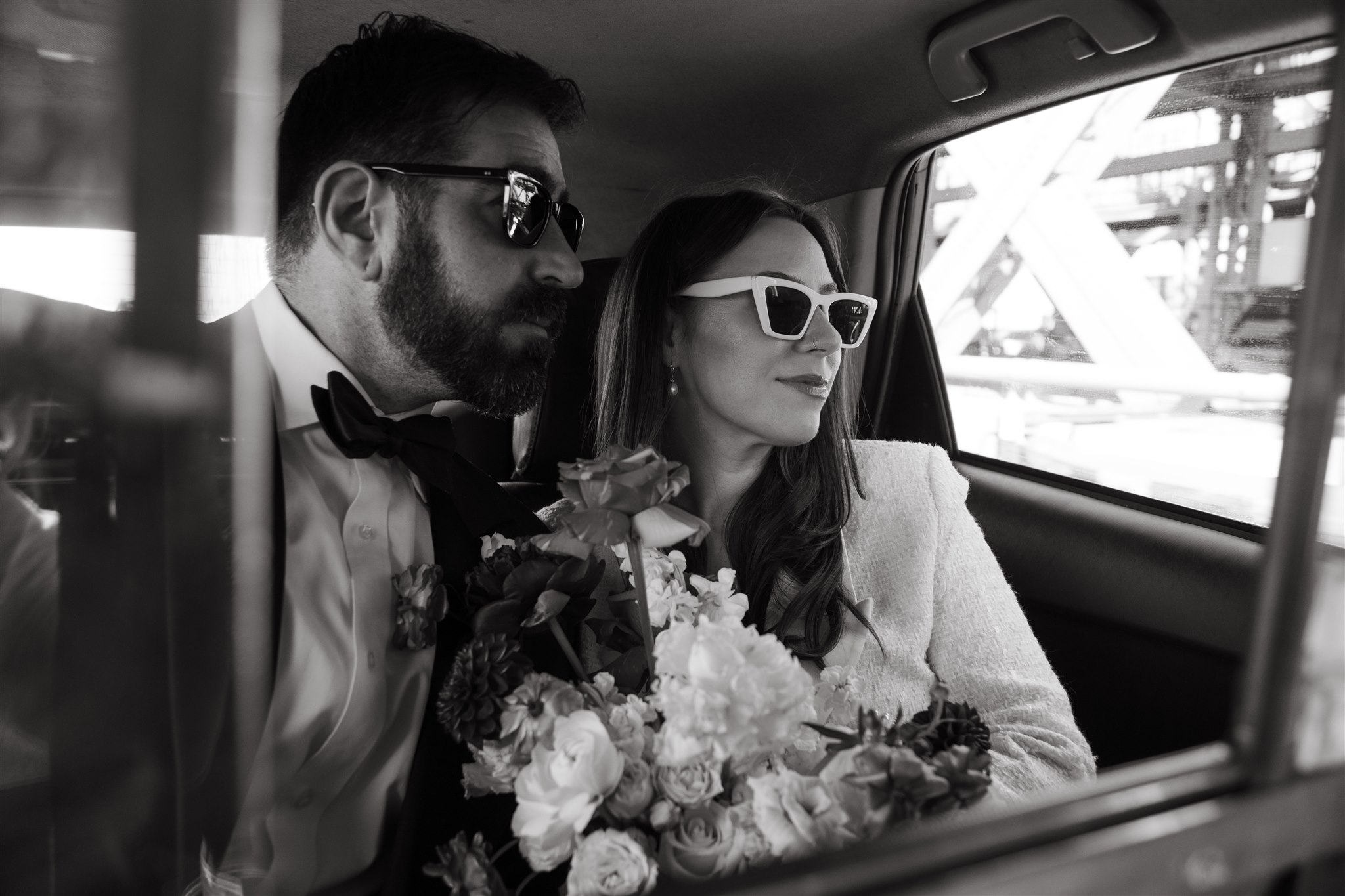 A couple in formal attire with sunglasses sitting in the backseat of a car, the woman holding a bouquet.