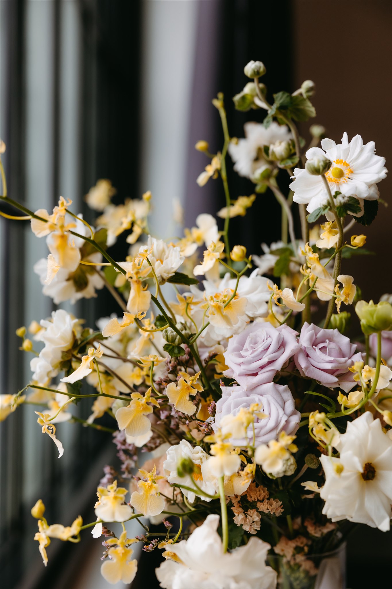A vibrant floral arrangement featuring pale purple roses, yellow orchids, and white daisies set against a softly blurred background.