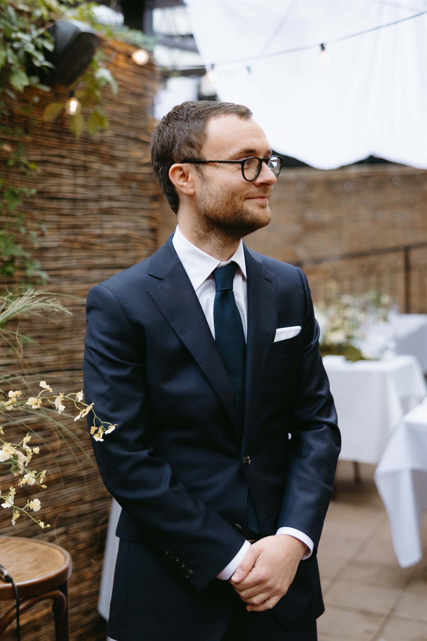 A man in a dark suit and tie with glasses smiles gently while standing in a decorated outdoor event space.