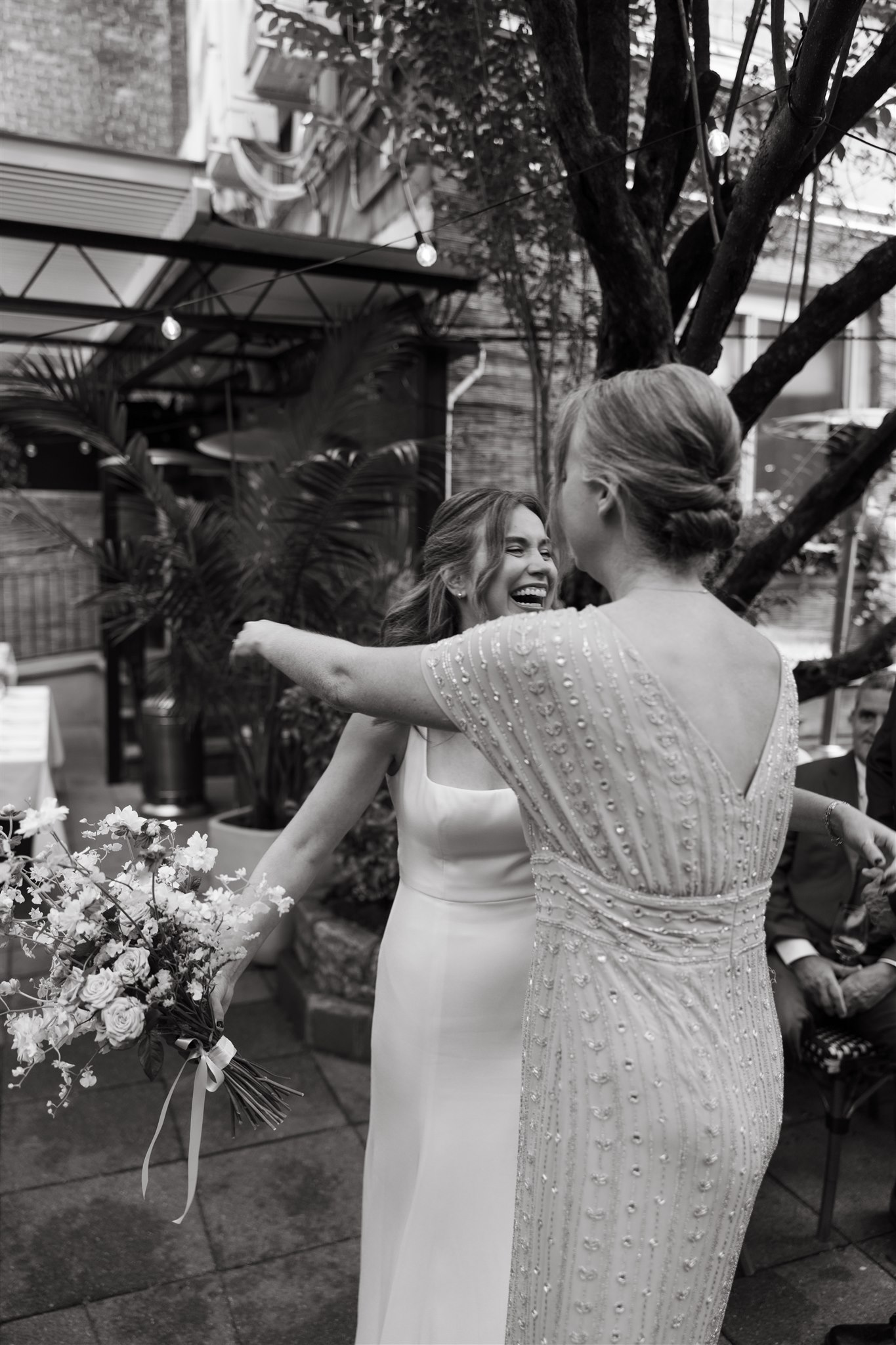 Two women, one in a white wedding dress and the other in a beaded dress, joyfully greet each other at an outdoor wedding, surrounded by guests and greenery.