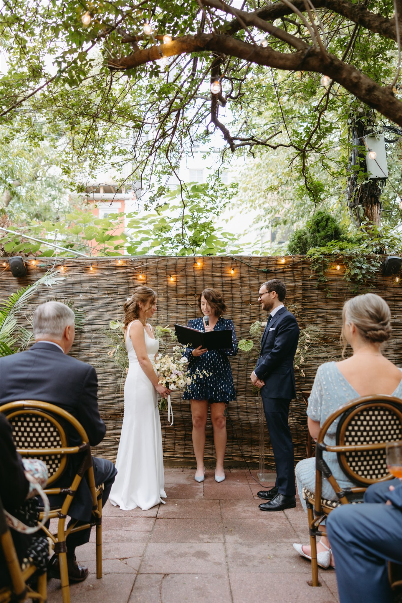 A bride and groom smiling during a wedding ceremony led by an officiant holding a black book, against a backdrop of string lights and a wicker screen at their restaurant wedding