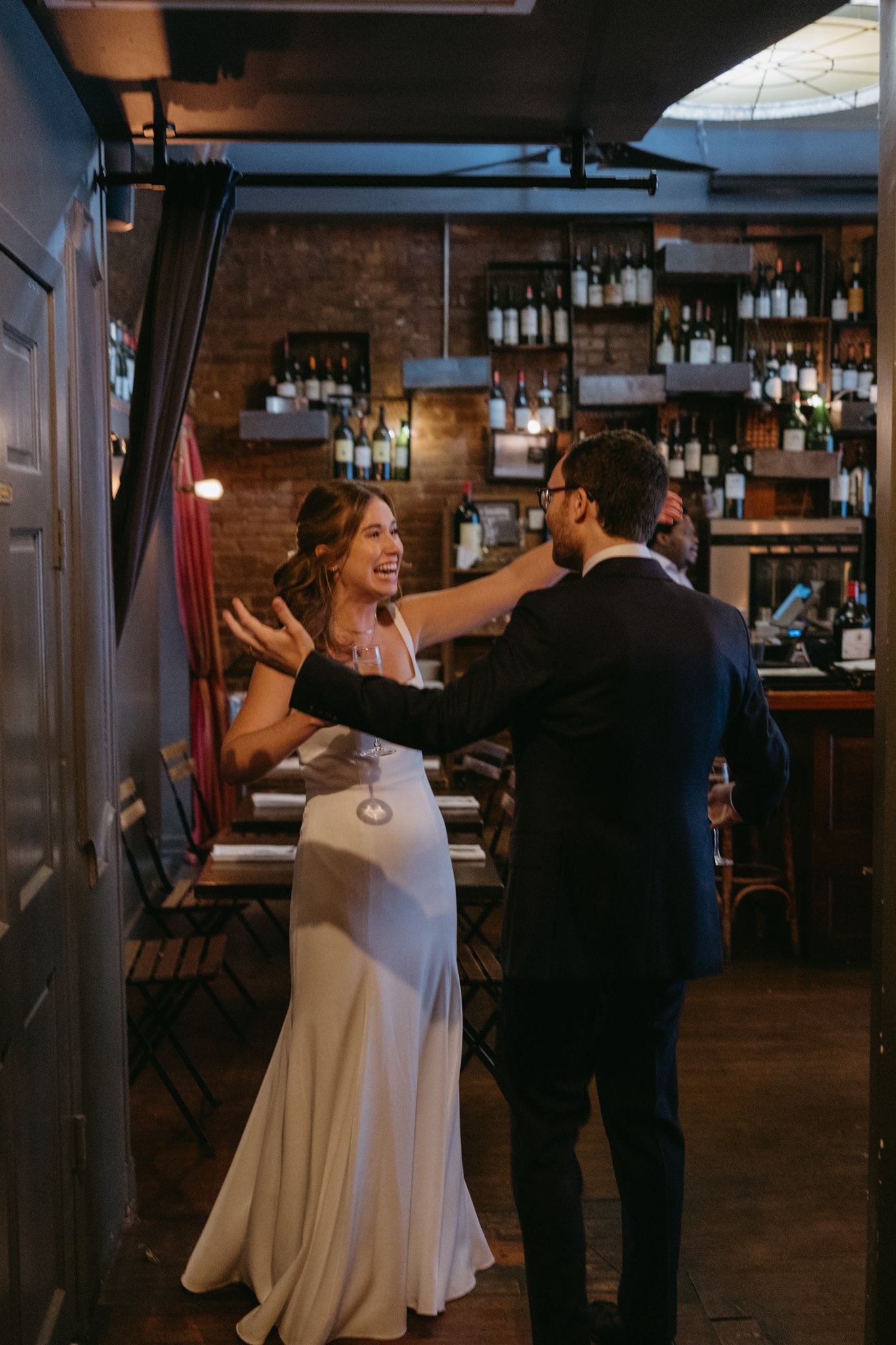 A couple happily dancing in a cozy bar with rows of bottles in the background. the woman is in a white dress, and the man is in a dark suit at their restaurant wedding