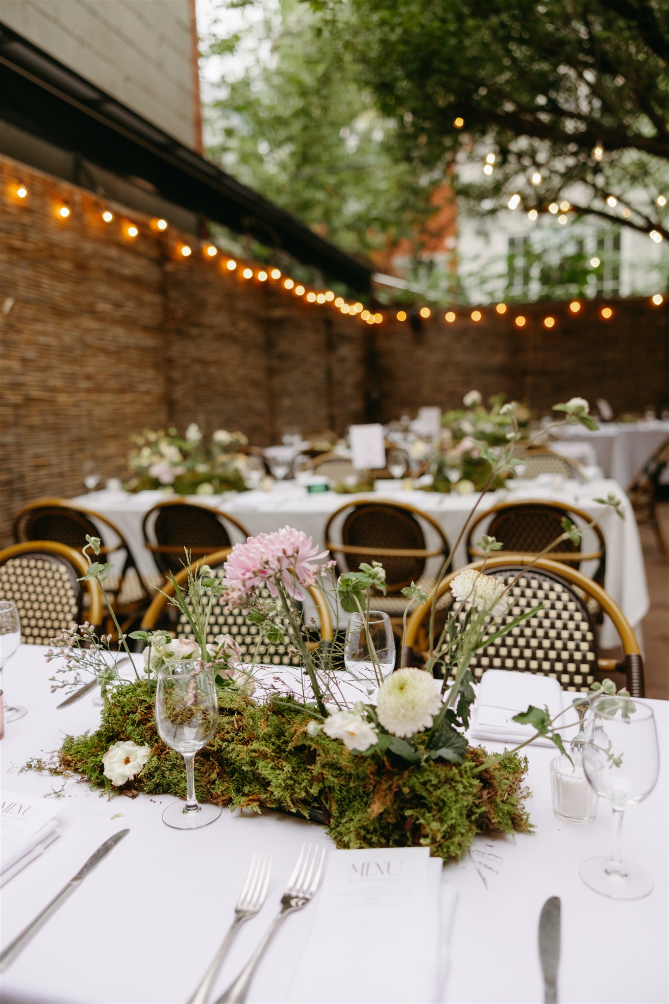 Outdoor dining area with wicker chairs and tables set with white tablecloths and floral centerpieces under string lights at an intimate restaurant wedding