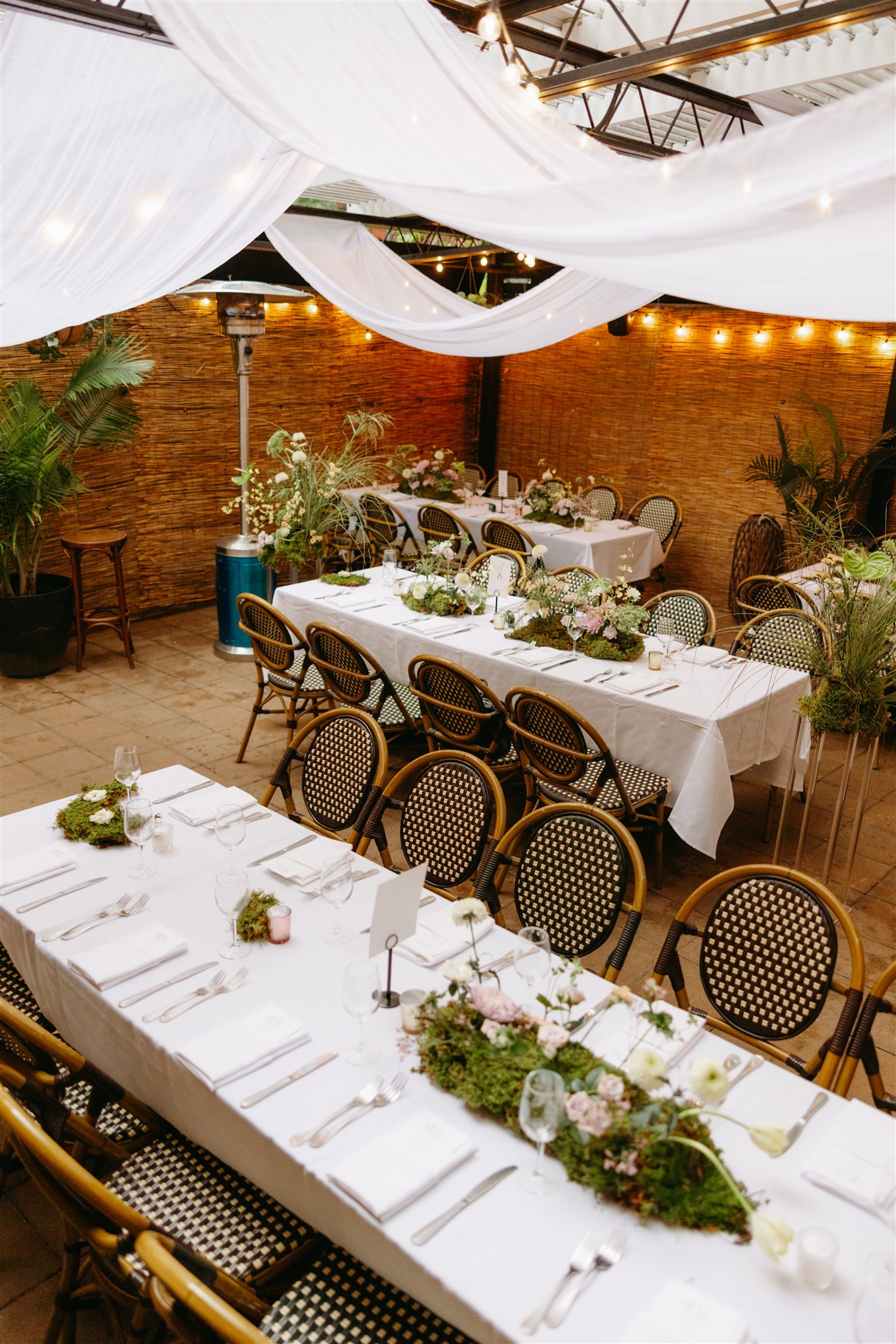 Outdoor dining area with wicker chairs and tables set with white tablecloths and floral centerpieces under string lights at an intimate restaurant wedding