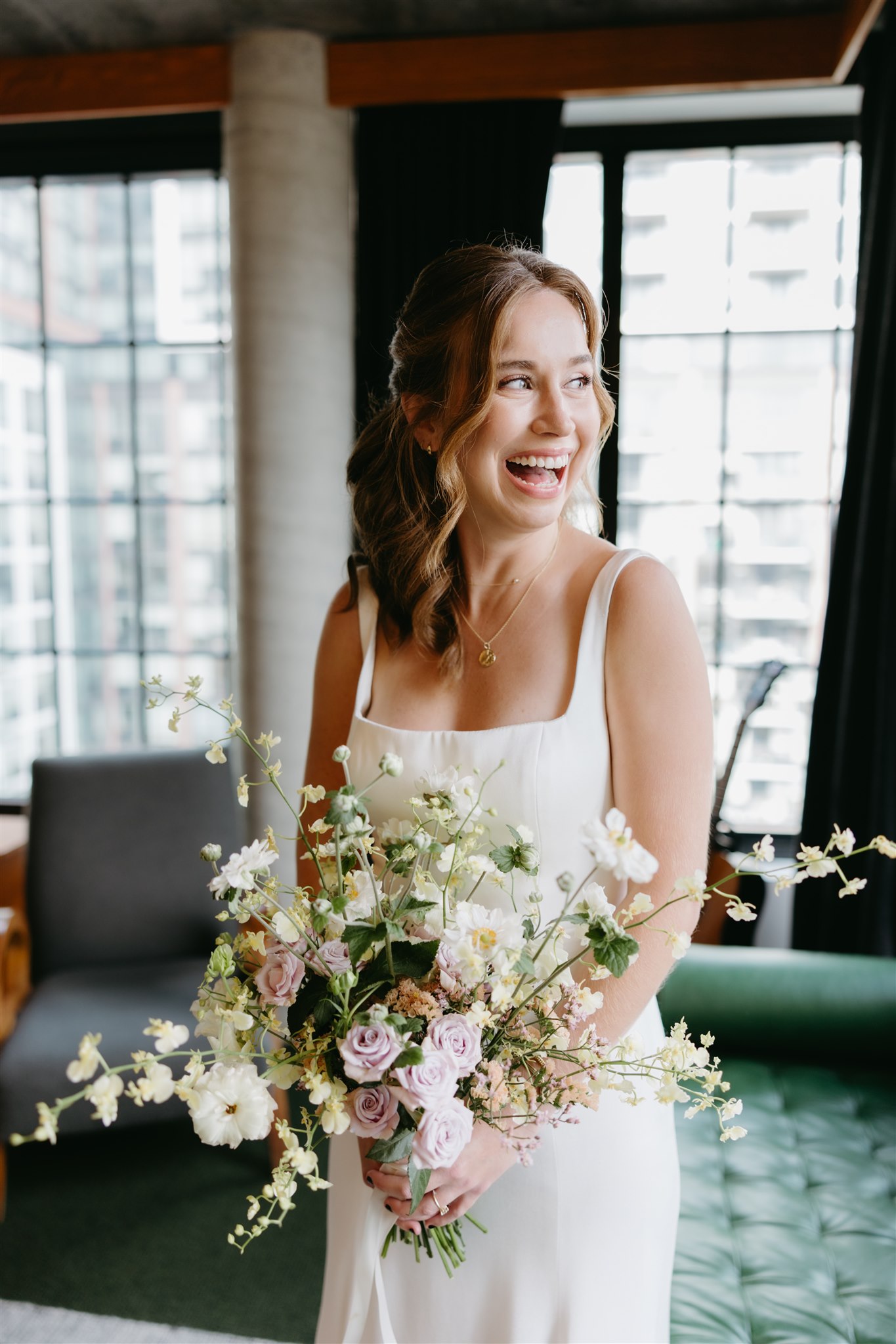 A joyful bride in a white dress holding a bouquet of pink and white flowers, laughing indoors with large windows in the background.