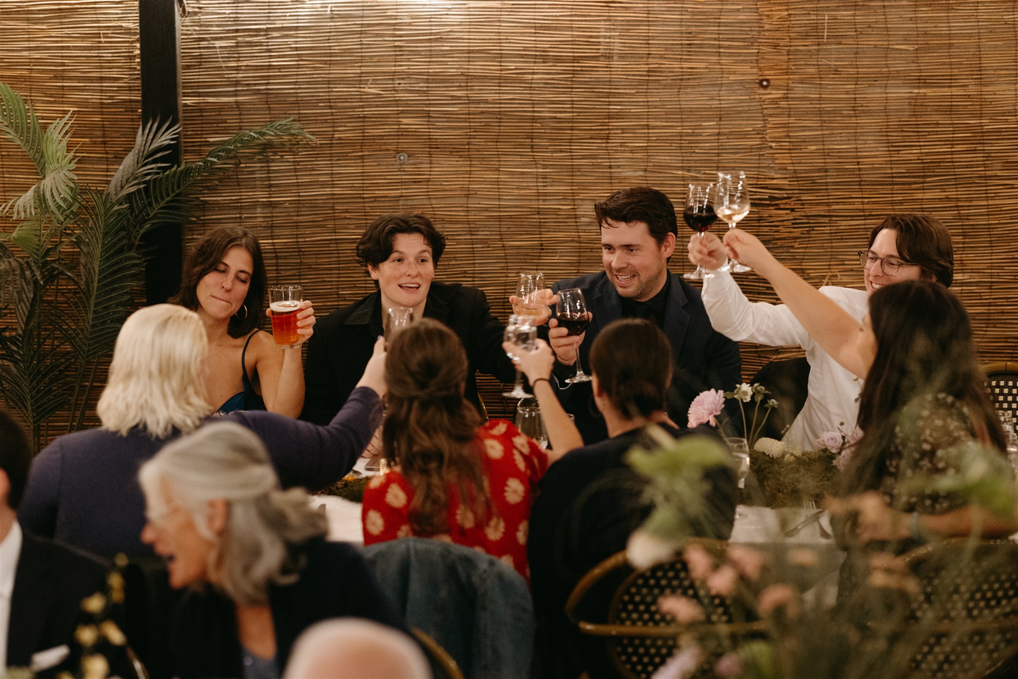 Group of friends toasting with wine glasses at a warmly lit dinner table, laughing and enjoying the evening at an intimate restaurant wedding