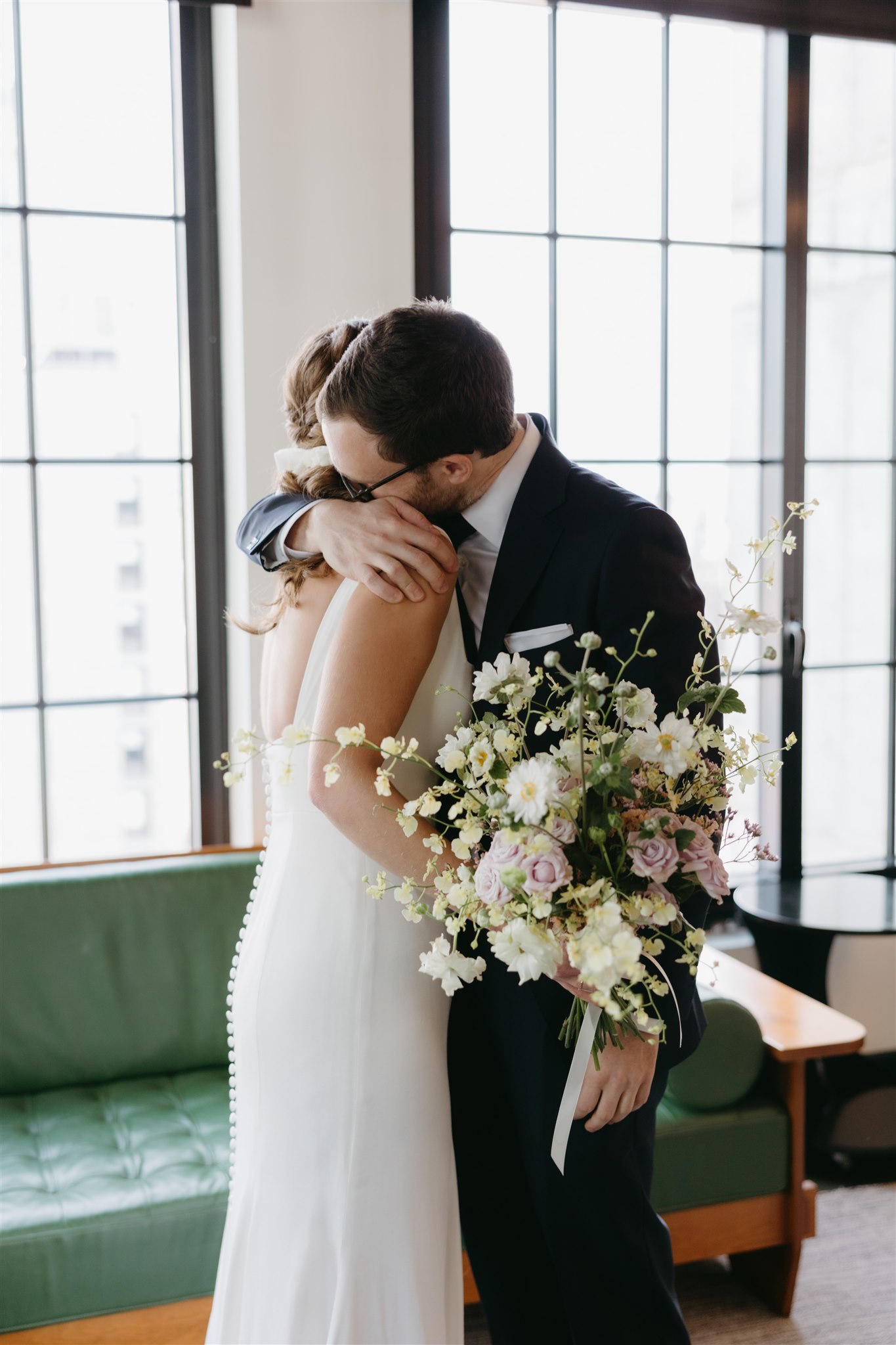 A bride and groom embracing, with the groom kissing the bride's forehead. the bride holds a bouquet of flowers, indoors by large windows.