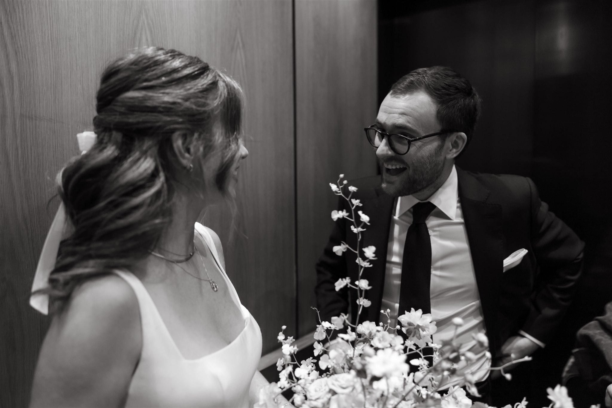 A bride and groom smiling at each other inside an elevator, with the bride holding a bouquet of flowers.