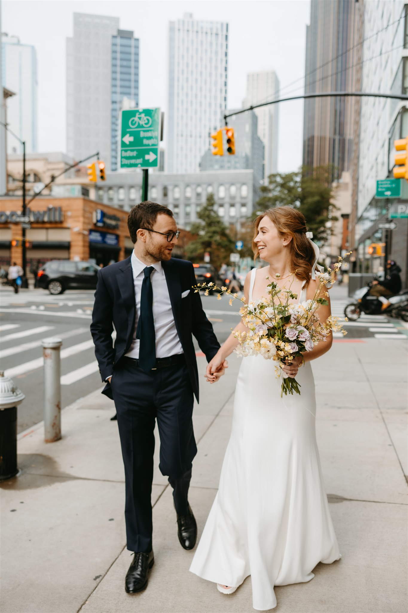 A bride and groom smiling at each other while walking across a city street, the bride holding a bouquet, and city buildings in the background.
