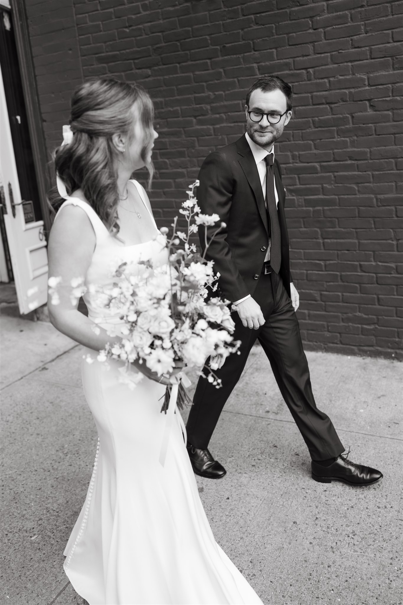 A bride and groom smiling at each other while walking across a city street, the bride holding a bouquet, and city buildings in the background.