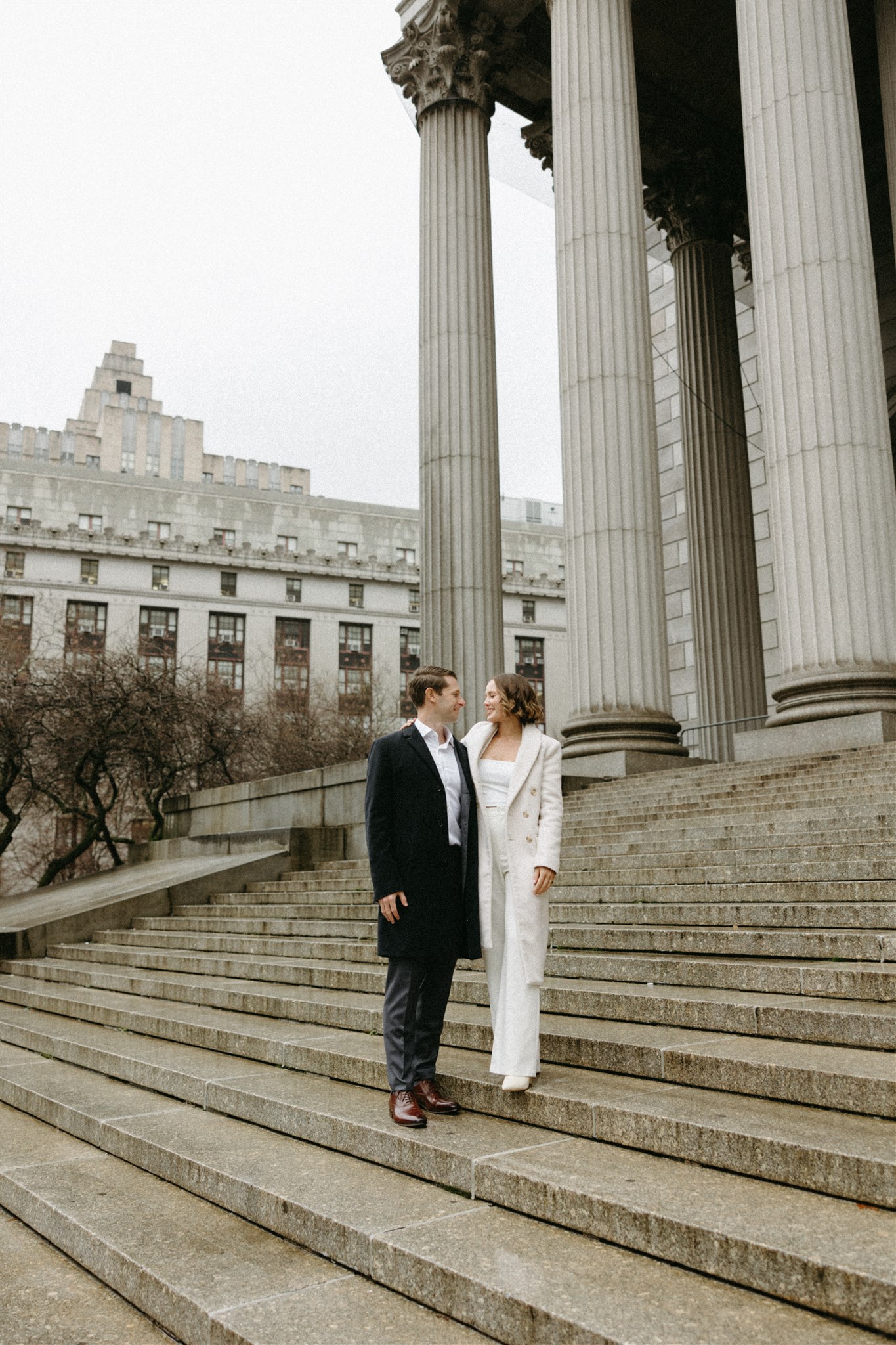 A couple stands on steps by neoclassical columns, facing each other and smiling as they elope in nyc
